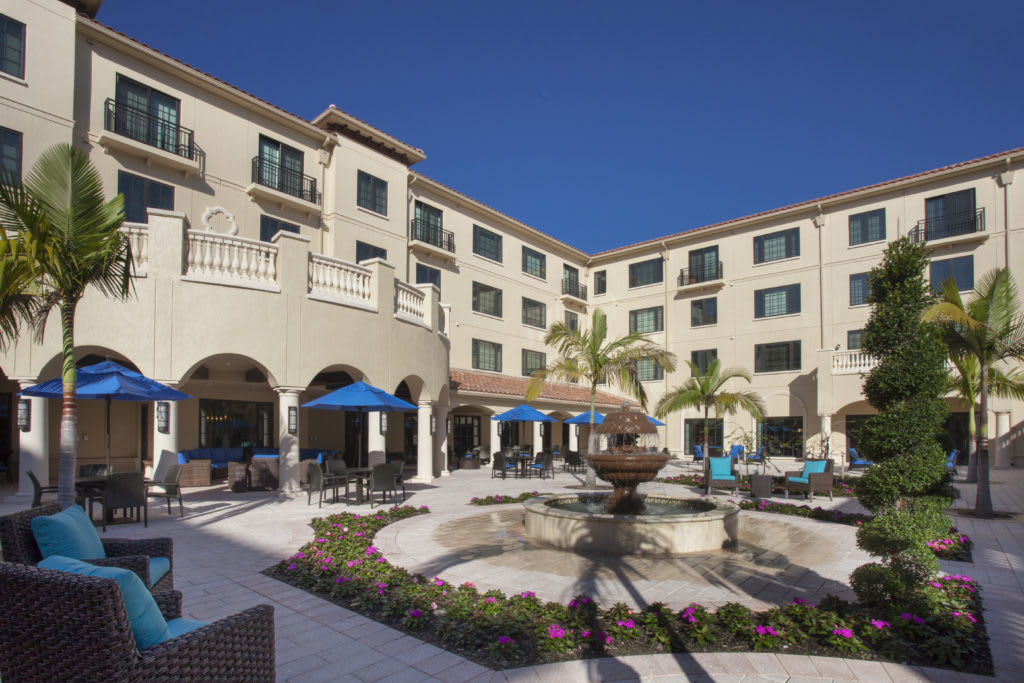A hotel courtyard featuring a central fountain, outdoor seating, and palm trees under a clear blue sky.