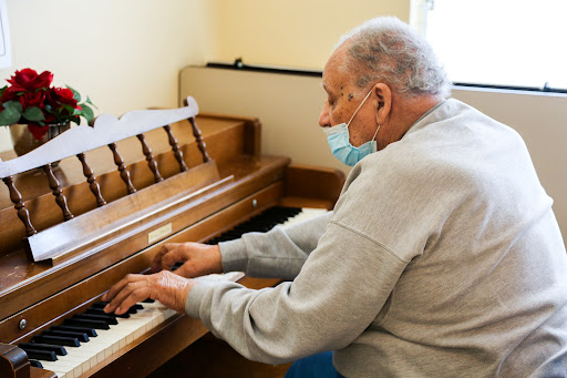 An elderly man playing piano indoors while wearing a mask.