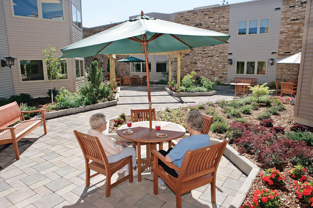 Two individuals sit at a wooden table with an umbrella on a sunny patio surrounded by a garden.