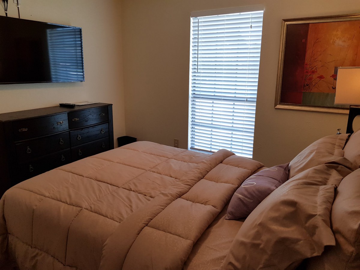 A cozy bedroom with a neatly made bed, dresser, wall-mounted TV, and a window with blinds.