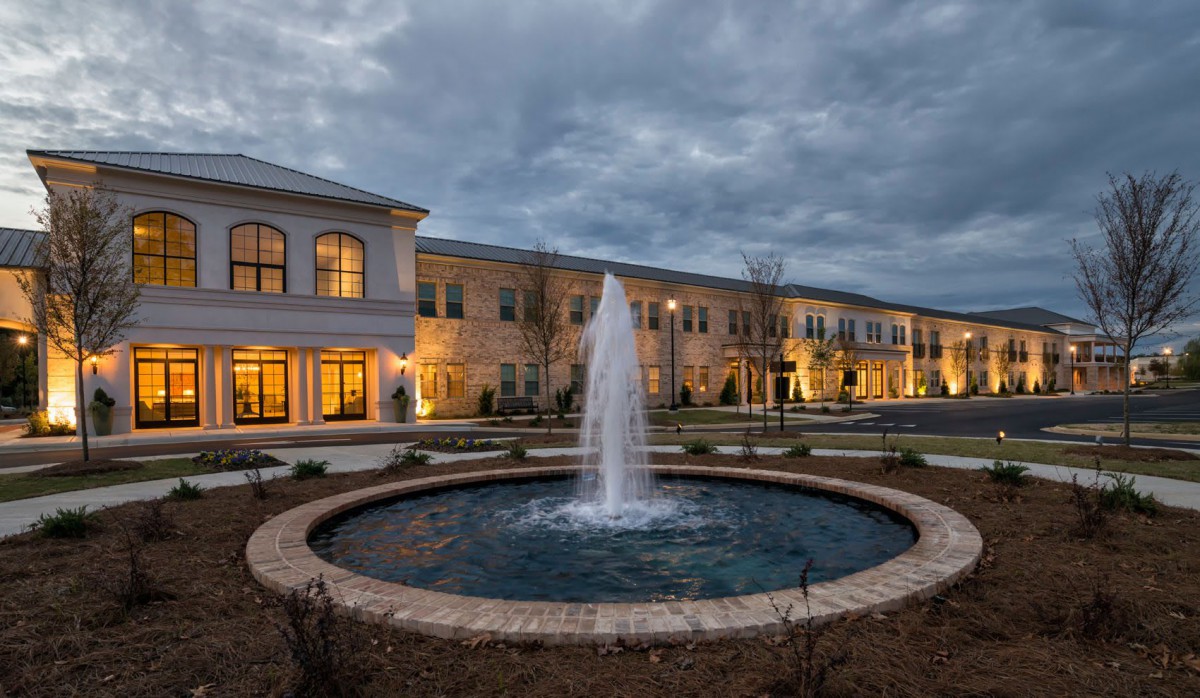 A tranquil scene of a building with illuminated windows and a central fountain under a cloudy evening sky.