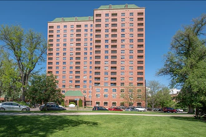 A tall residential apartment building with multiple floors, surrounded by trees and parked cars under a clear blue sky.