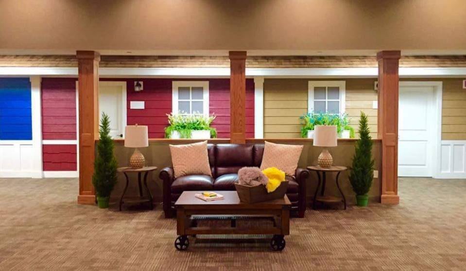 A cozy living room setup with a leather sofa, decorative pillows, and elegant lamps flanking a wooden coffee table. The backdrop features window-like decor with potted plants.