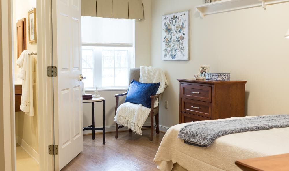 A cozy bedroom with a chair, pillow, and blanket next to a window letting in natural sunlight.