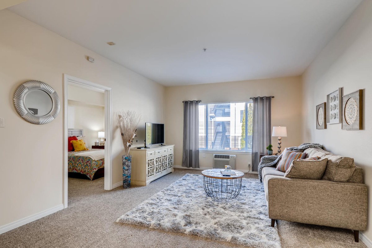 A cozy living room with modern decor, featuring a sofa, coffee table, and TV. The space is well-lit by a large window with curtains, and there's an open doorway leading to a bedroom.