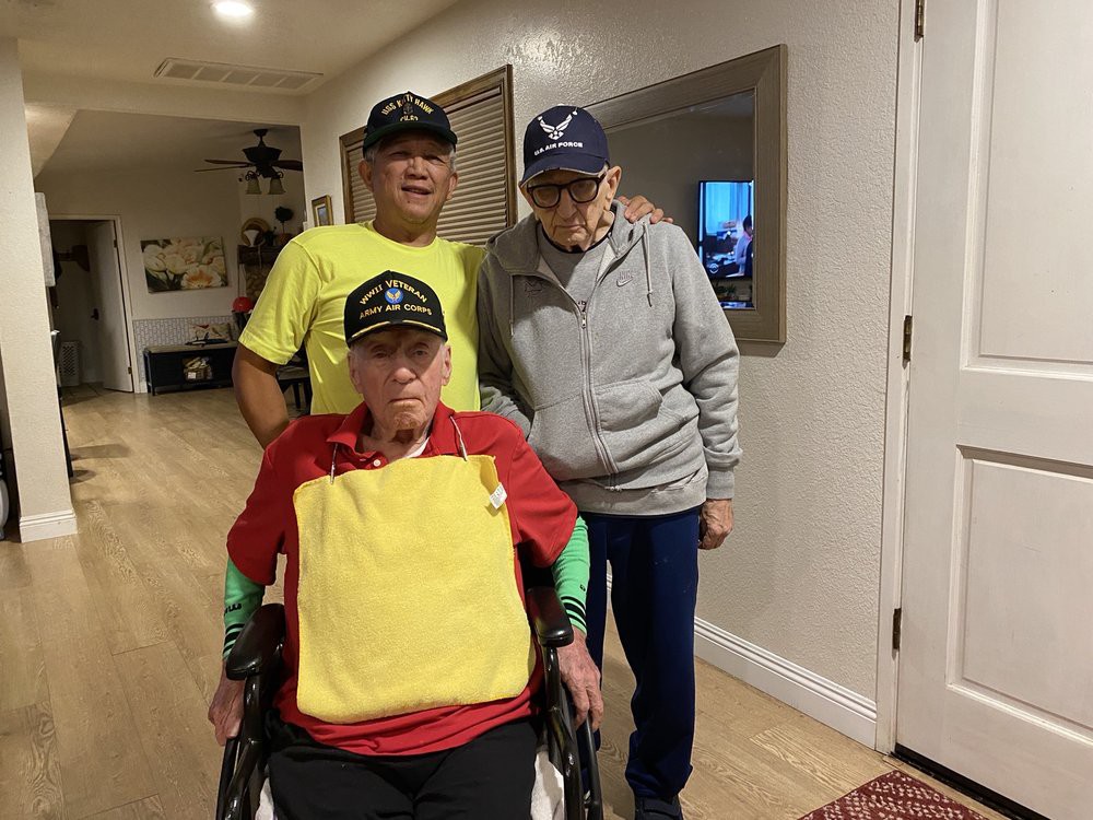 Three elderly individuals, two standing and one in a wheelchair, pose together wearing hats inside a home.