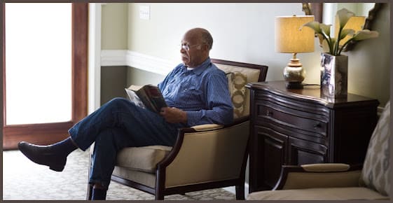 A man sitting in a chair reading a book beside a lamp.