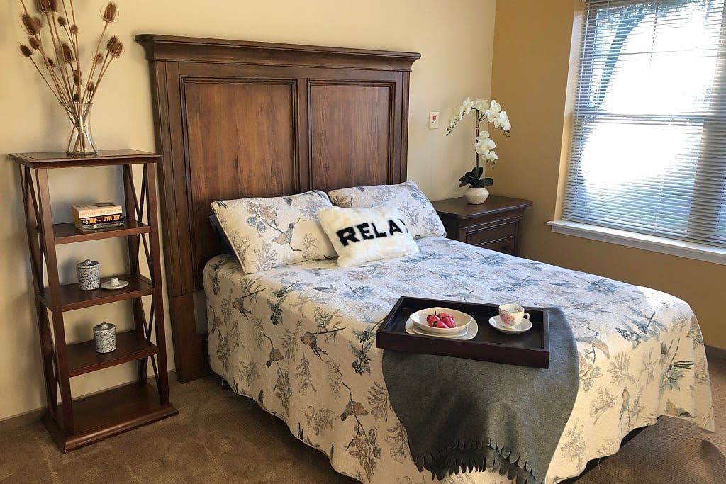 A cozy bedroom with a neatly made bed, featuring floral bedding and a 'RELAX' pillow. A tray with breakfast is placed at the foot of the bed, and an orchid decorates the nightstand next to a window.