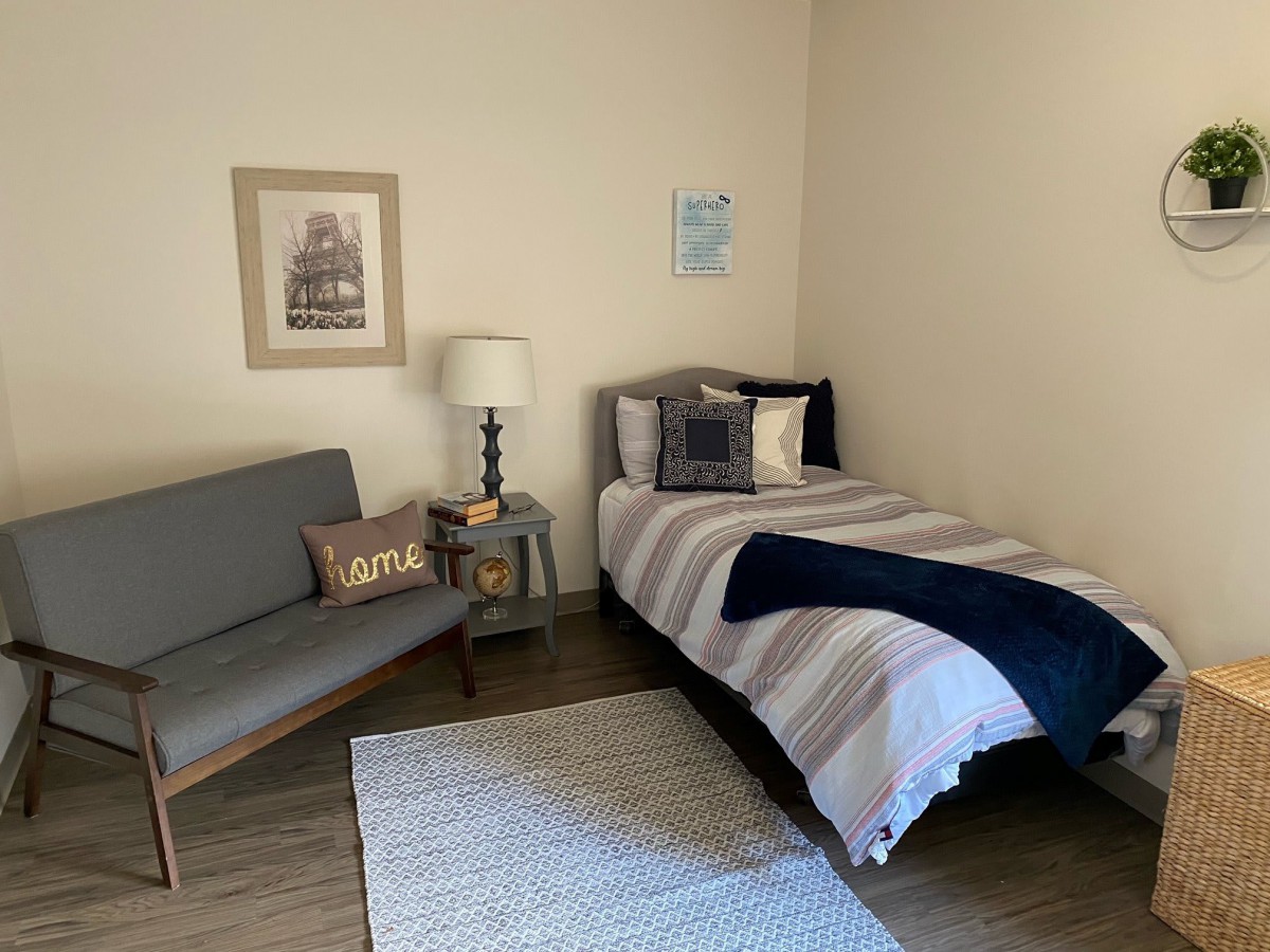 A minimalist bedroom featuring a single bed with striped bedding, a small sofa, a table lamp, and simple wall decor.