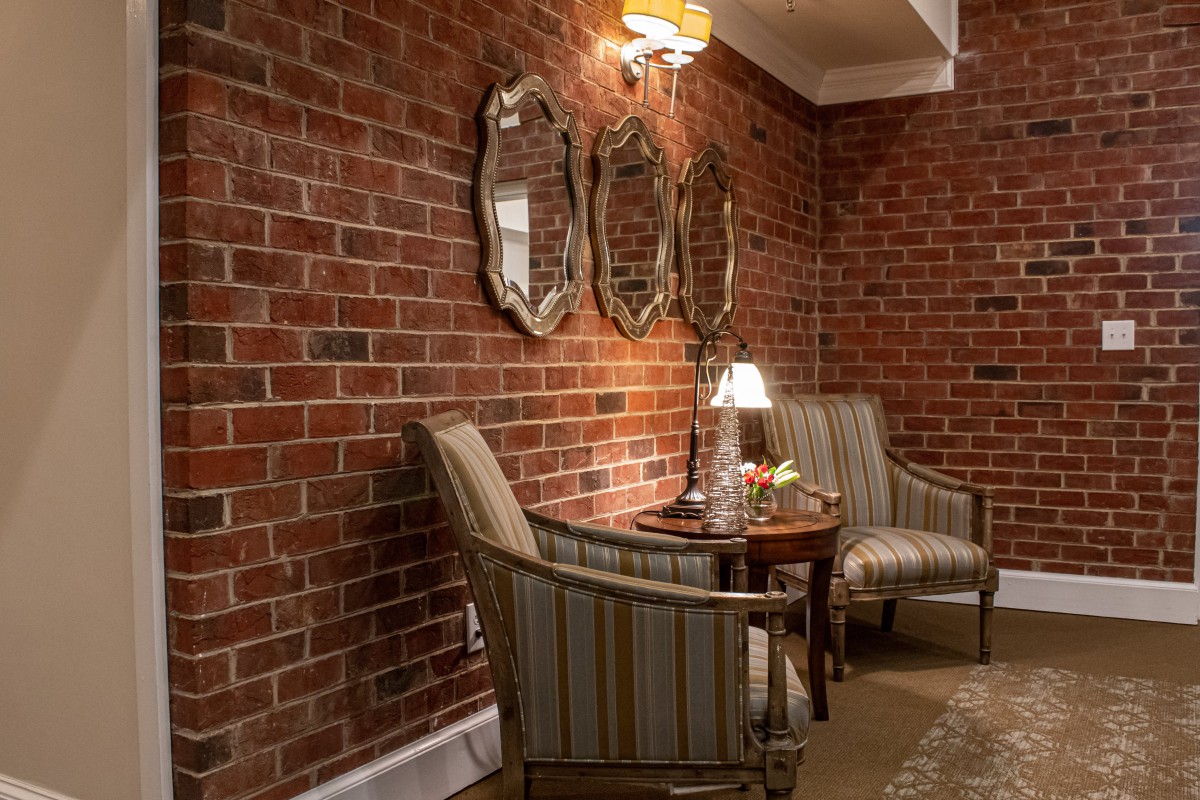 A cozy seating area with armchairs and a table against a brick wall, adorned with mirrors and illuminated by a lamp.