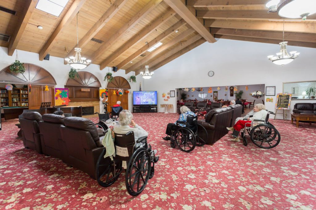 A cozy communal living room in a nursing home, featuring residents in wheelchairs watching television.