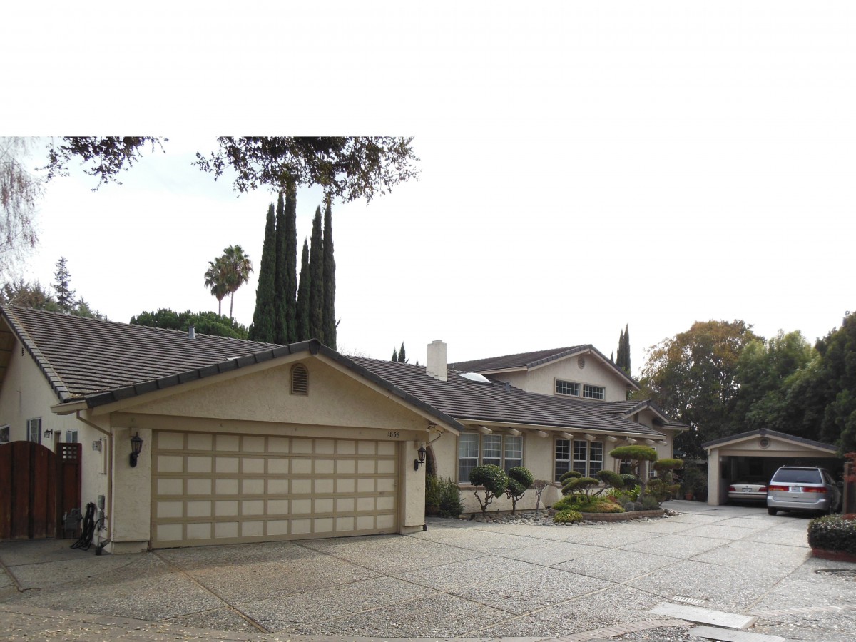 A suburban residential house with a large driveway and attached garage, surrounded by tall trees.