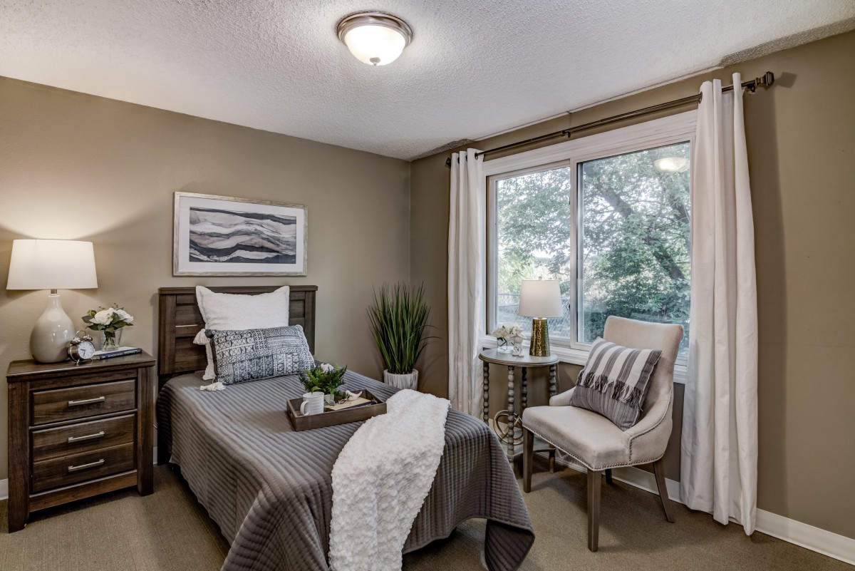 A cozy bedroom featuring a single bed with gray bedding, a large window allowing natural light, and elegant decor including a chair, lamp, and decorative plants.