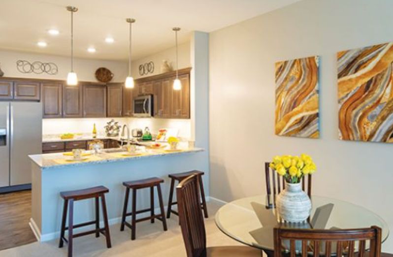 A modern kitchen and dining area featuring wooden cabinetry, pendant lighting, bar stools, and a glass dining table with a vase of yellow flowers.