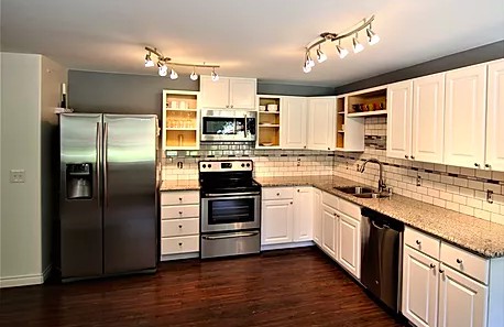 A modern kitchen with stainless steel appliances and white cabinetry, featuring hardwood floors and track lighting.