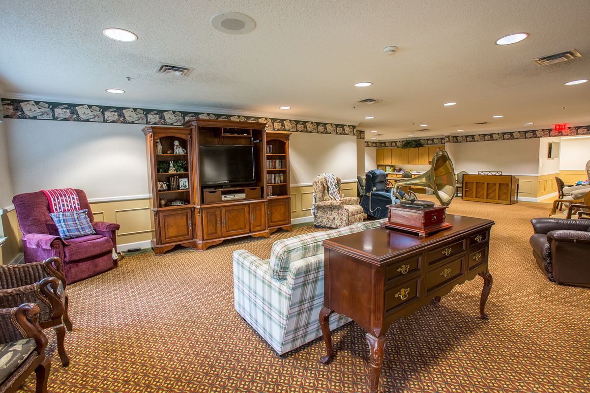 A spacious living room featuring a TV, bookshelf, and comfortable seating with wooden furniture on a carpeted floor.
