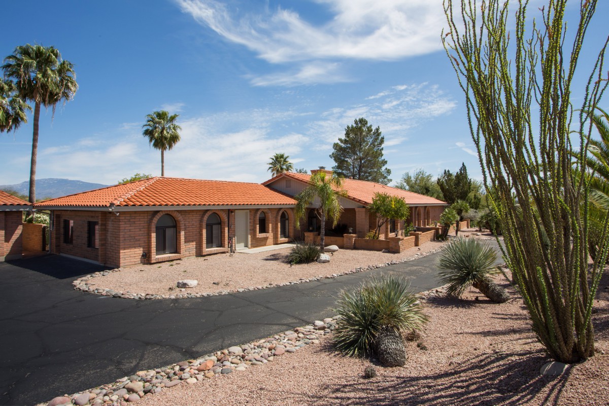 A desert-style house with a red tile roof, surrounded by a dry landscape and palm trees under a clear blue sky.