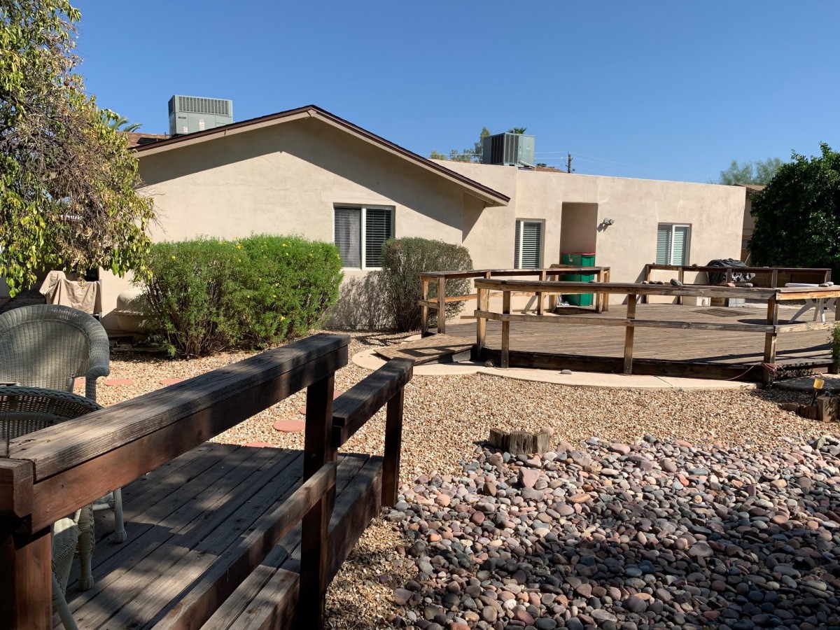 A single-story house with a wooden ramp, surrounded by desert landscaping and gravel.