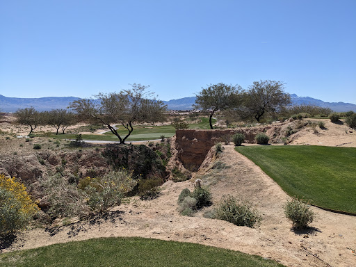 A scenic view of a golf course in a desert setting, featuring green grass, trees, and mountains in the background.
