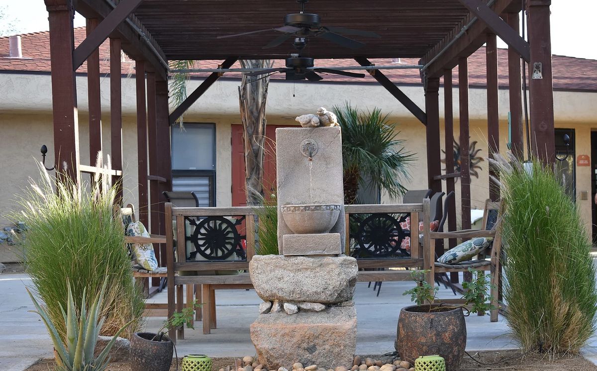 A stone fountain under a wooden gazebo surrounded by plants in an outdoor setting.