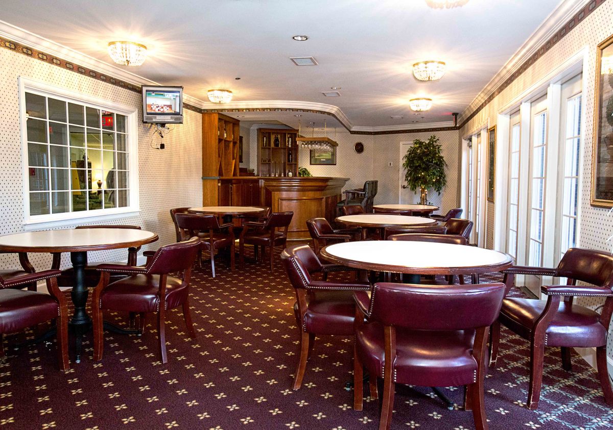 A cozy dining area with round tables and upholstered chairs, featuring carpeted flooring and large windows.