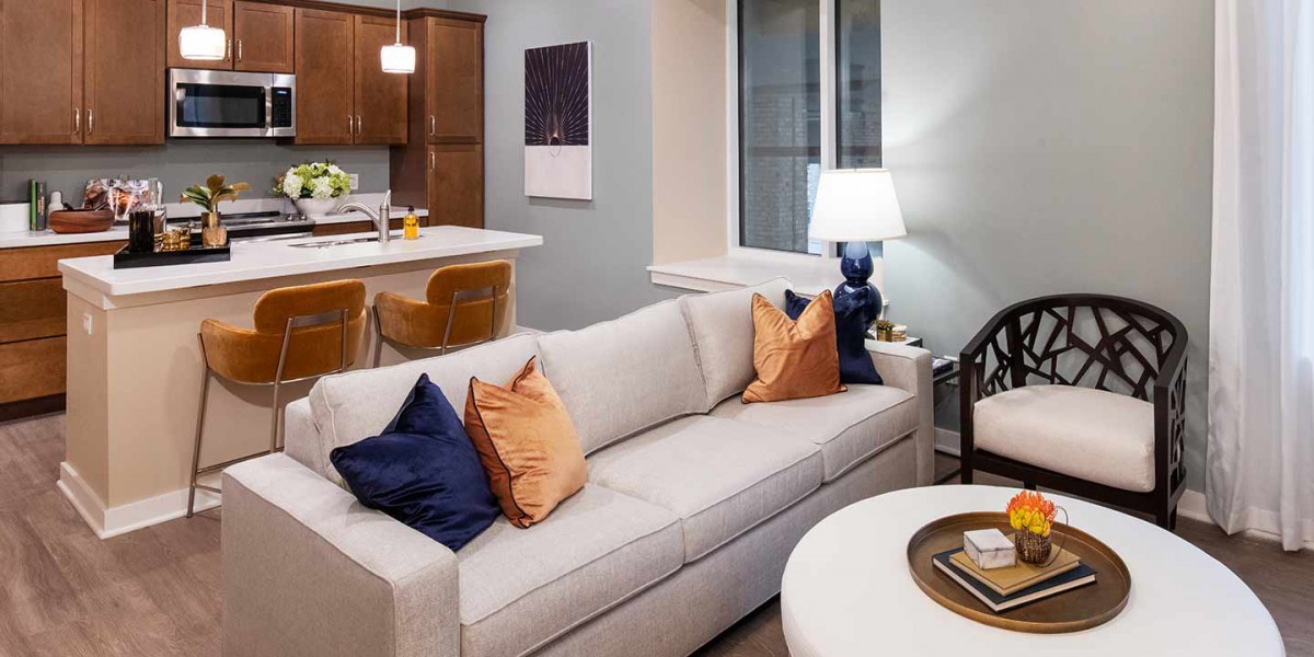 A cozy living room with a light gray sofa adorned with orange and dark blue pillows, adjacent to an open kitchen.