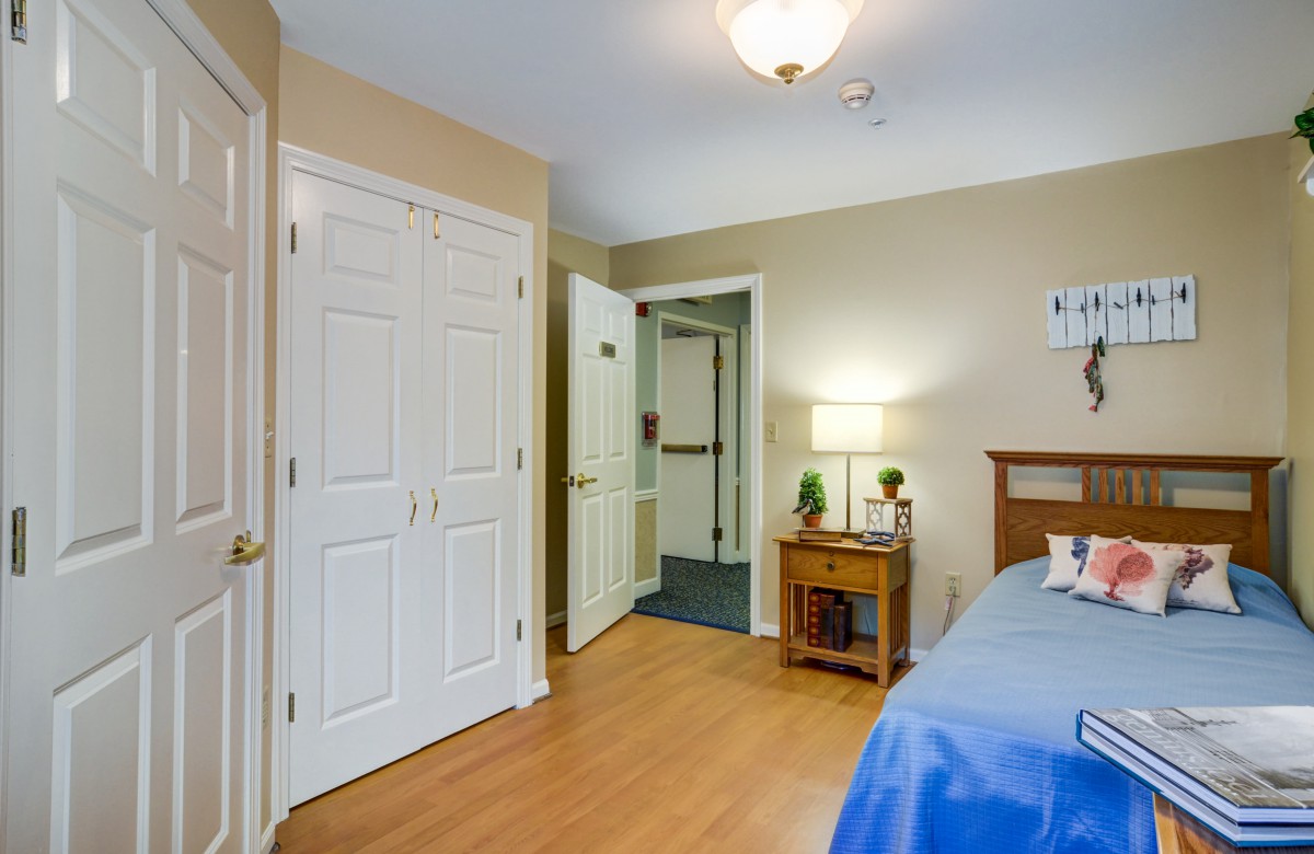 Cozy bedroom interior with wooden flooring, featuring a closet and a nightstand with a lamp.