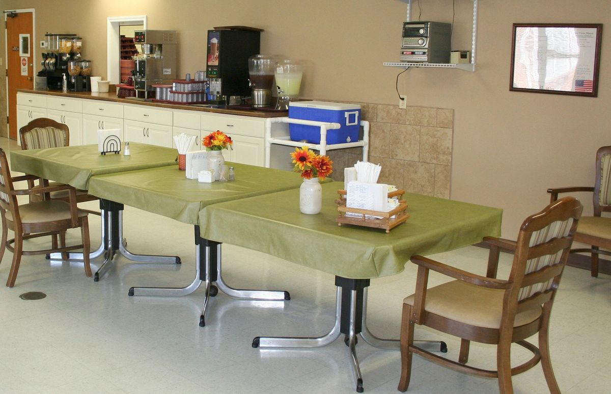 A room with tables covered in green tablecloths and decorated with flower arrangements, alongside a kitchen area.