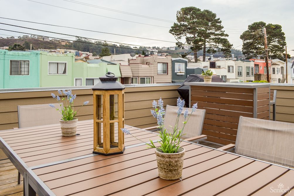Cozy rooftop patio with a wooden table, potted plants, and a lantern overlooking an urban neighborhood.