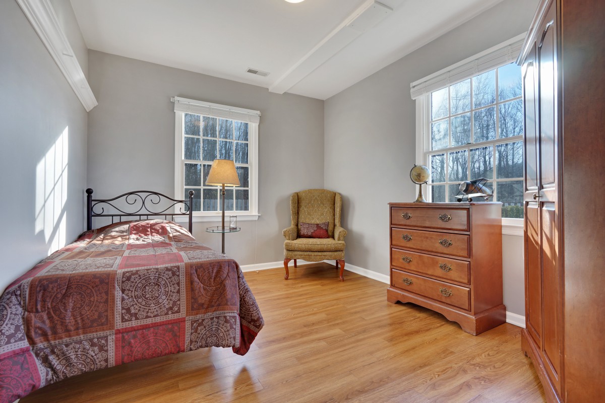 A cozy bedroom featuring a single bed, armchair, dresser, and ample natural light from two windows.