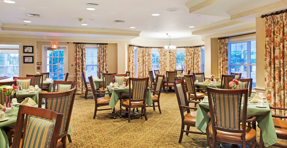 A well-lit dining area with neatly arranged tables and chairs, featuring elegant curtains and carpeting.