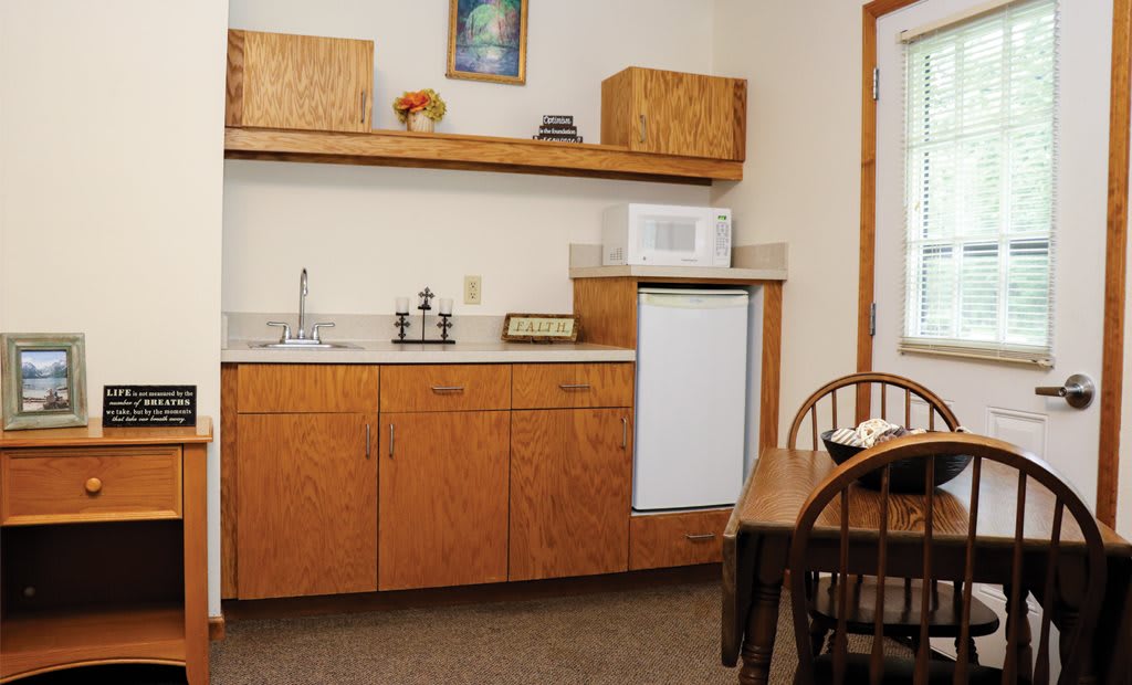 A compact kitchenette with wooden cabinets, a microwave, and a small refrigerator next to a dining table with chairs.
