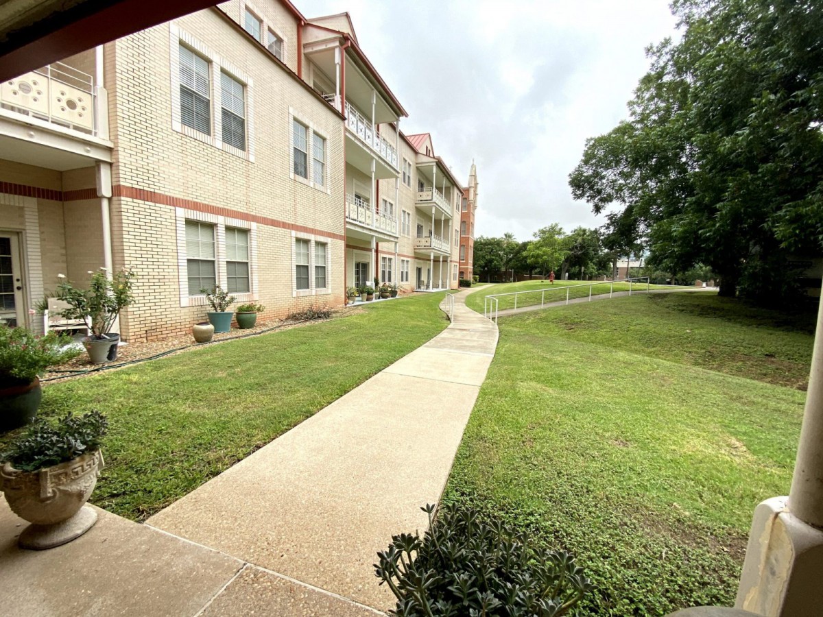 A sidewalk leads to a multi-story apartment building surrounded by a well-maintained lawn and trees on a cloudy day.