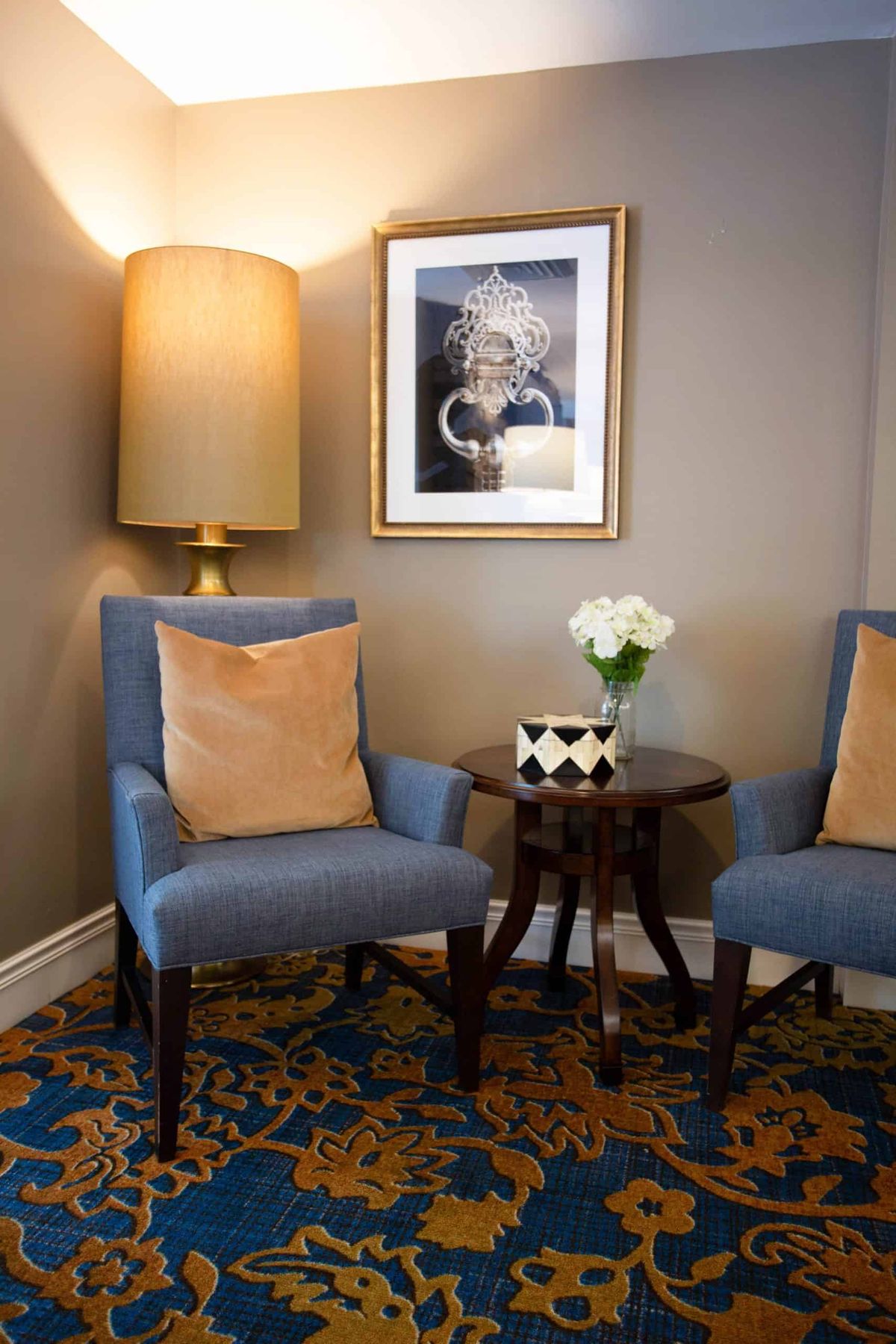 A cozy seating area with two blue chairs, a small round table with flowers, a large lamp, and framed artwork on the wall.