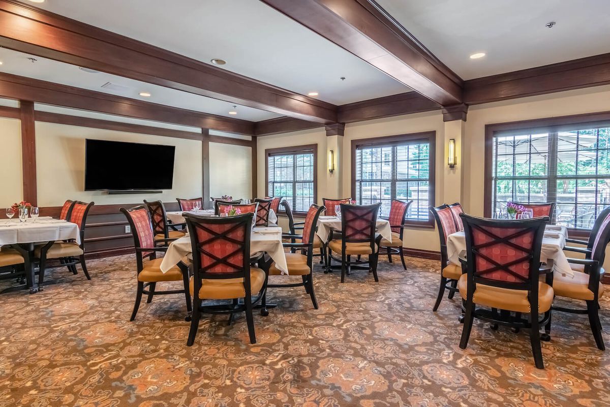 A cozy dining room with multiple tables and chairs, featuring large windows and a television on the wall.