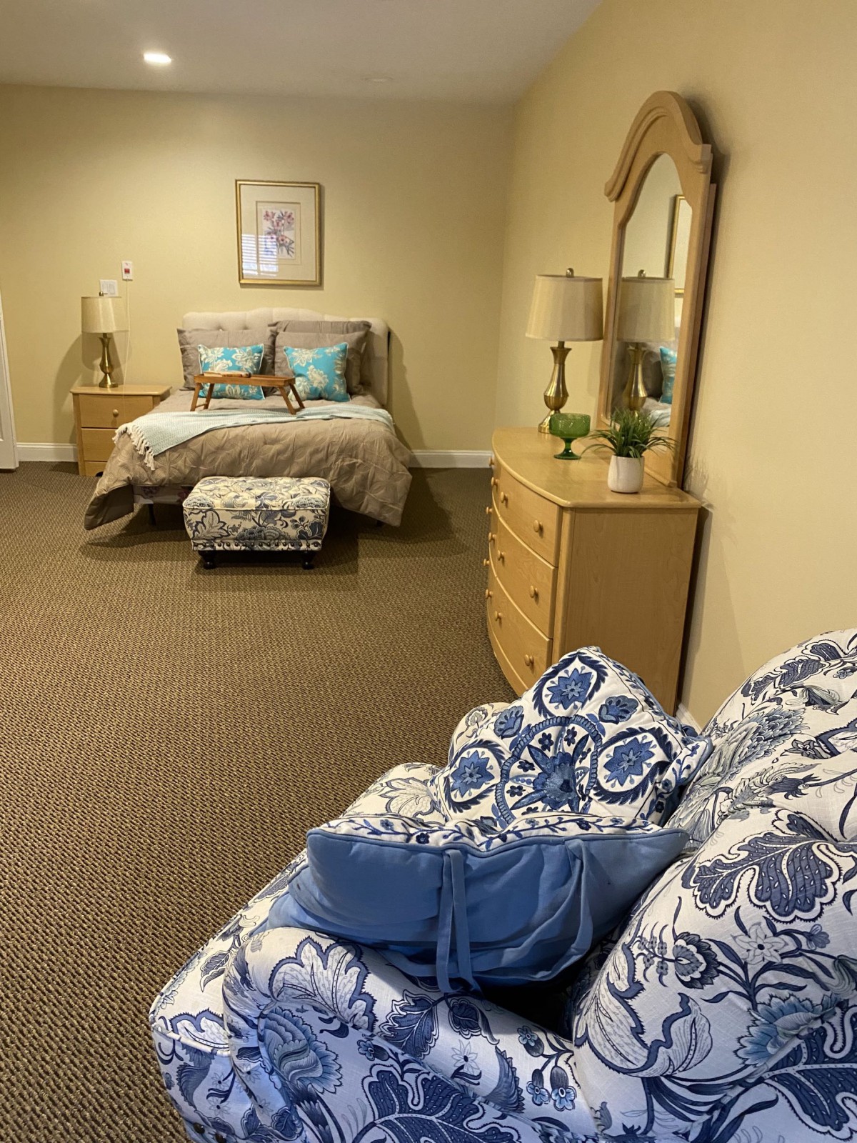 A cozy bedroom featuring a bed with patterned cushions, a dresser with a mirror, and warm lighting.