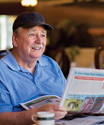 A man in a hat is smiling while reading a newspaper indoors.