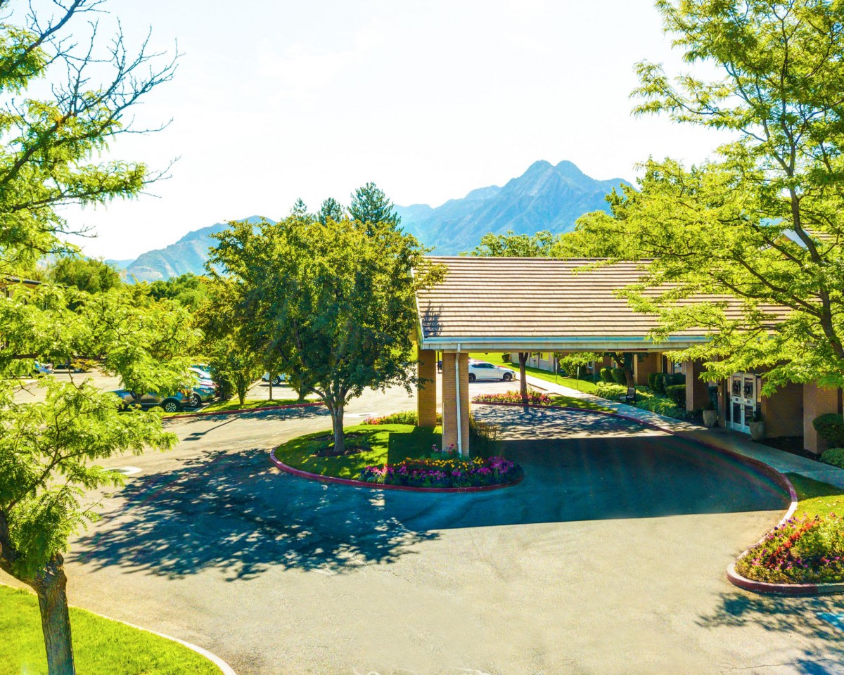 A scenic view of a building entrance surrounded by trees, with mountains in the background on a sunny day.