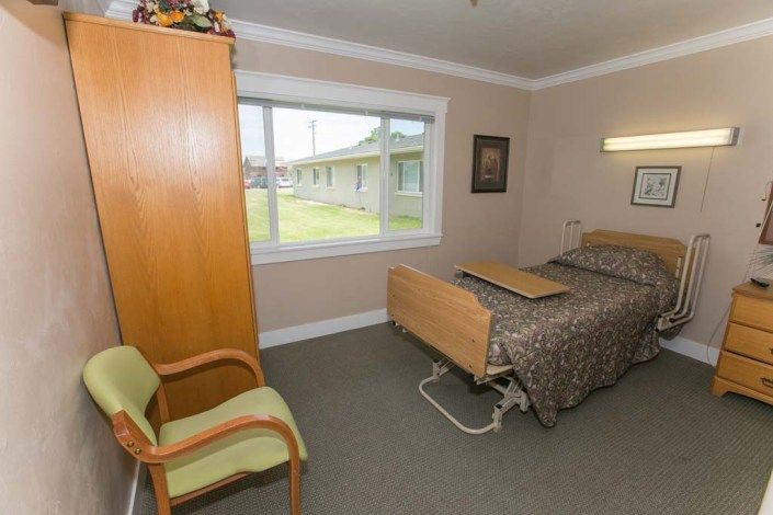 A simple nursing home room with a single bed, wooden wardrobe, and a large window providing natural light.