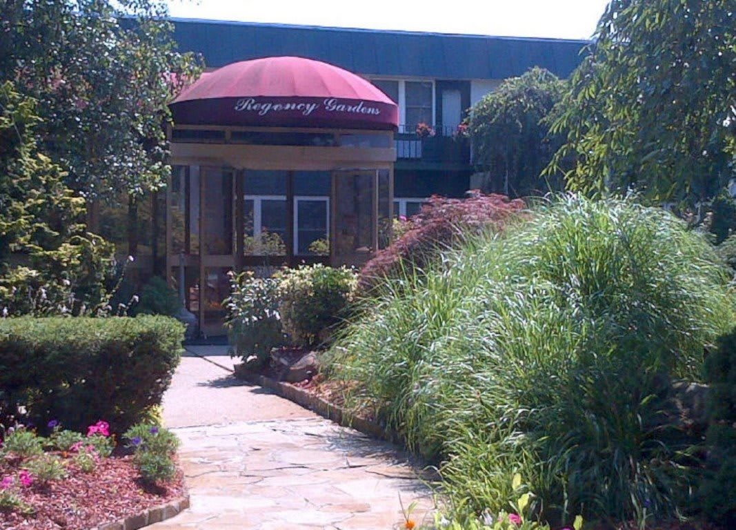 A building entrance with a maroon canopy, surrounded by lush greenery and a stone pathway.