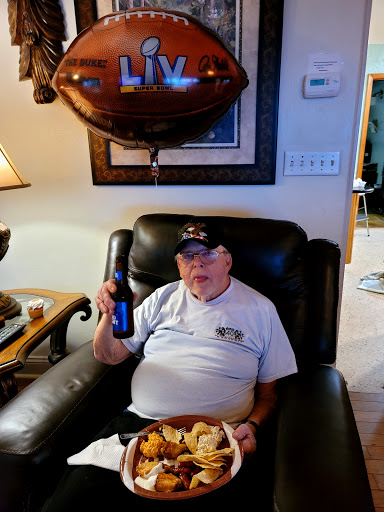 An elderly man enjoying snacks and a drink while relaxing in his recliner, celebrating with a festive atmosphere.