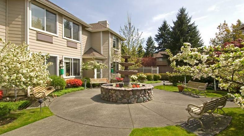 A peaceful courtyard with a central stone fountain surrounded by lush greenery and flowers, adjacent to a residential building.