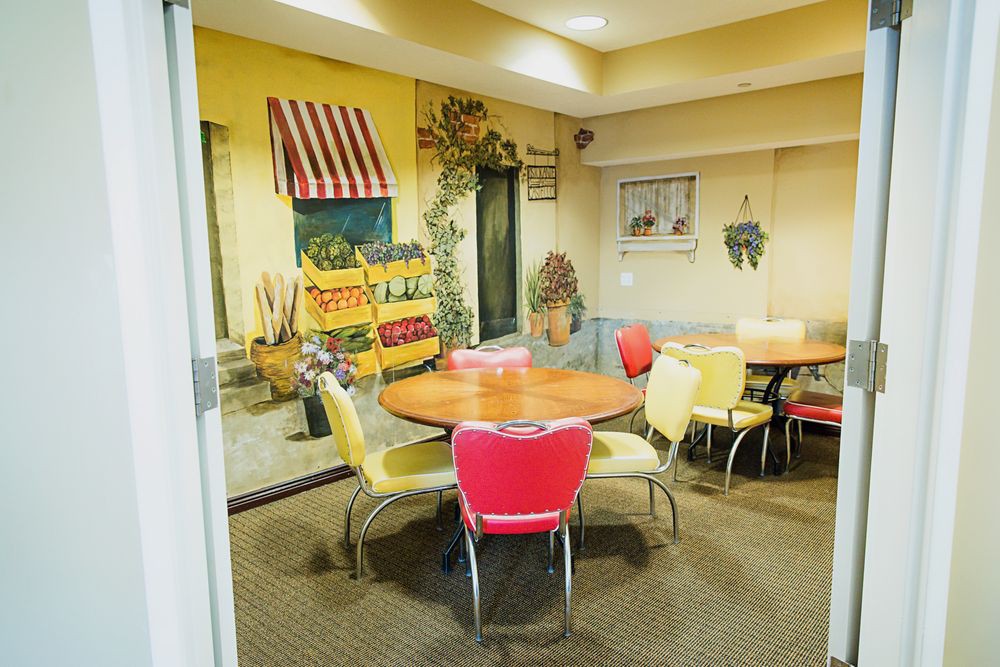 A cozy indoor dining area with round tables and colorful chairs, featuring a retro-style wall mural depicting an outdoor market scene.