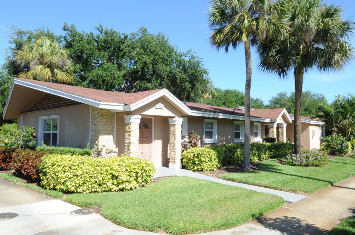 A suburban bungalow surrounded by a neatly manicured lawn and palm trees, set against a clear blue sky.