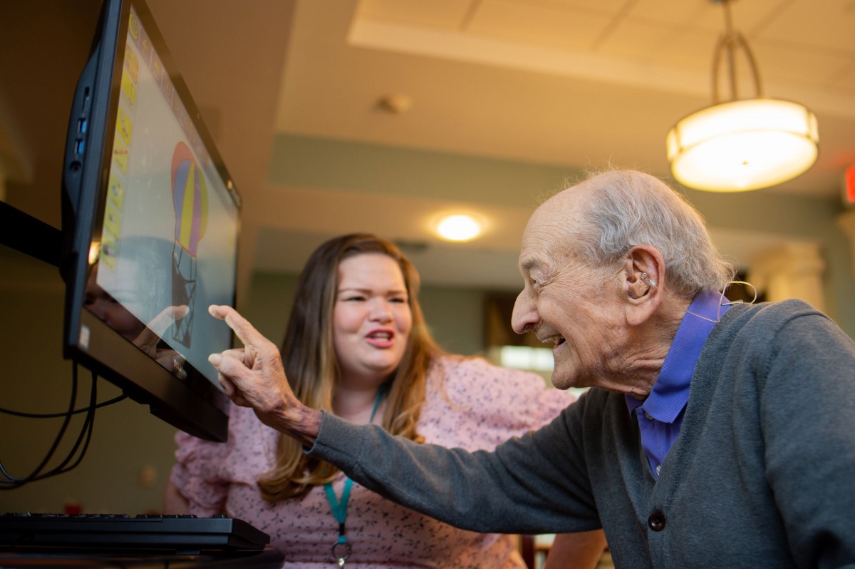 An elderly man and a young woman smiling and interacting with a computer screen.
