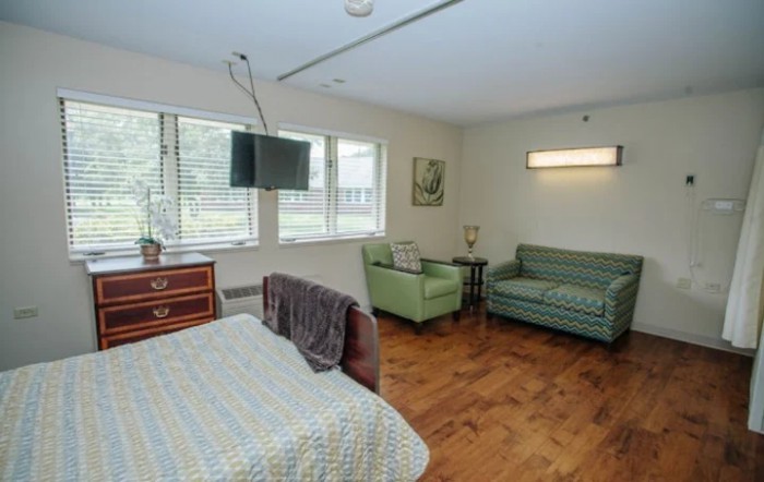 A hospital room with a bed, armchair, and sofa, featuring a wall-mounted television and large windows.