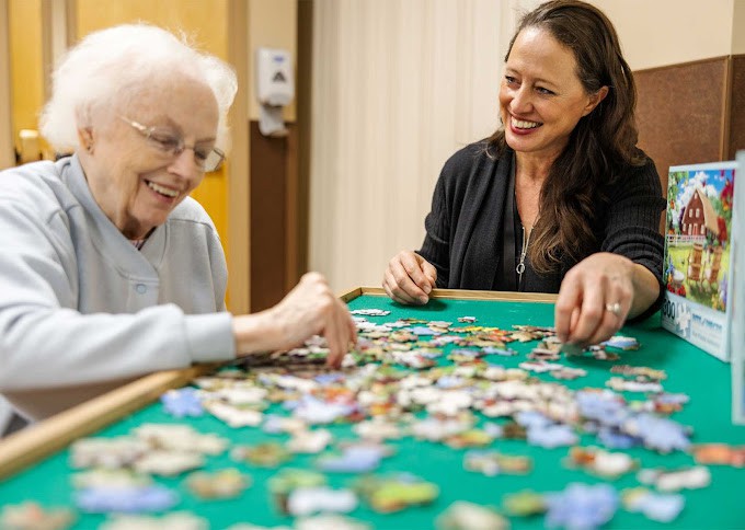 An elderly woman and a caregiver enjoy putting together a puzzle indoors.
