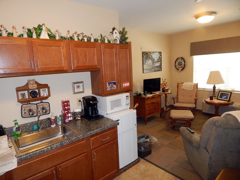 A small, cozy apartment kitchen with a living area in view, featuring wooden cabinets, a microwave, refrigerator, and various decorations.