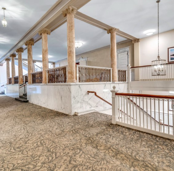 A spacious hallway with elegant columns and carpeted flooring, featuring a staircase with wooden railings.