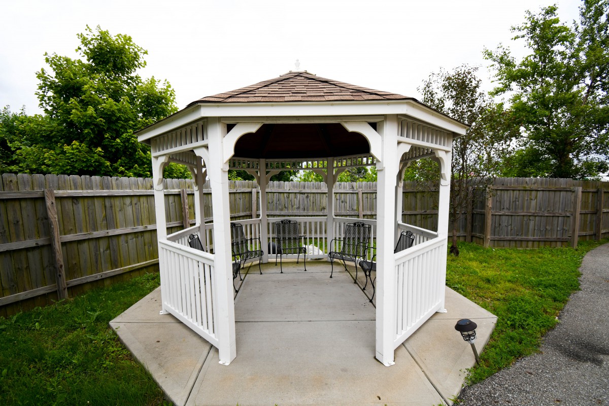 A white wooden gazebo in a fenced backyard surrounded by greenery.